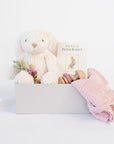 White Lulu Rabbit plush from Manhattan Toy Company, pink dried floral bundle, Peter Rabbit hardcover book, wooden teething rattle with pink silicone, light pink muslin swaddle. All in grey gifting box 