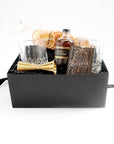 Black box with a set of two crystal tumblers, a double sided golden jigger, bottle of old fashioned syrup with black and gold label, a pack of playing cards  with elaborate gold and bronze foil design, pack of dried oranges.