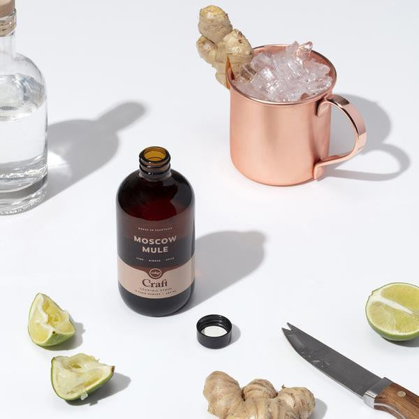 Open brown Moscow Mule bottle surrounded by limes, ginger root, a bar knife, a bottle of vodka and a copper Moscow mule mug with ice and a ginger garnish. 