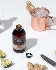 Open brown Moscow Mule bottle surrounded by limes, ginger root, a bar knife, a bottle of vodka and a copper Moscow mule mug with ice and a ginger garnish. 