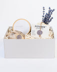 Light grey box containing Potager soap co. lavender soap wrapped in white paper closed with brown twine and purple "PS" wax seal. Lavender bath bomb wrapped in white tissue paper with floral filigree design and purple "PS" wax seal, dried lavender bundle, gold candle tin with white "lavender" sticker.