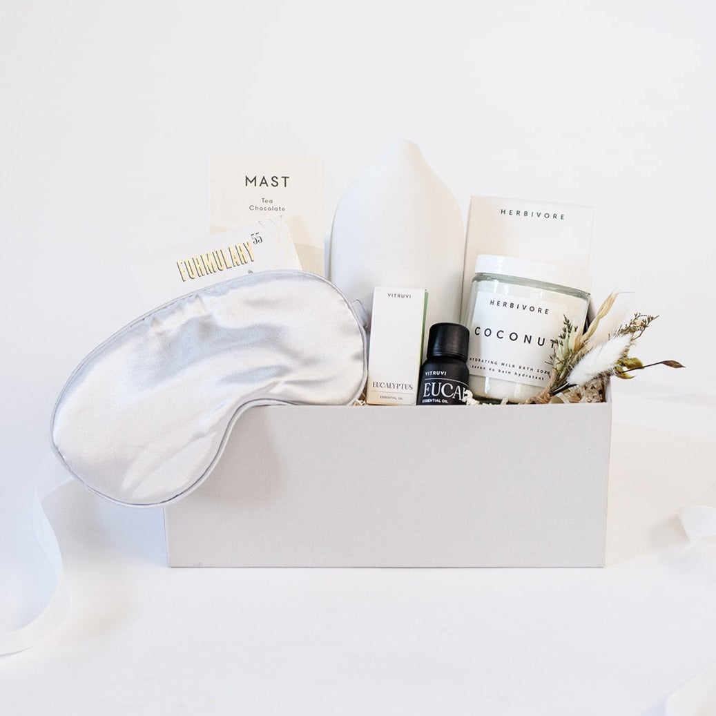 Light grey box containing an Ivory sleep mask, Formulary 55 Eucalyptus &amp; Peppermint foot soak, Mast tea chocolate bar, Vitruvi white stone diffuser, black bottle of Vitruvi eucalyptus oil next to it&#39;s white box, glass jar of Herbivore coconut bath soak next to it&#39;s white box, dried floral bundle. 