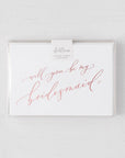 White envelope size card (approx. 3.5" x 5"), reads, "Will you be my bridesmaid?" in rose gold text in a clear plastic bag,