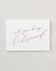 White envelope size card (approx. 3.5" x 5"), reads, "Will you be my bridesmaid?" in rose gold text.