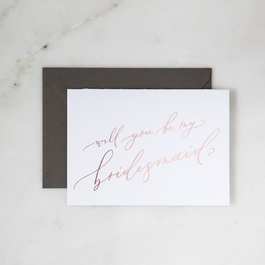 White envelope size card (approx. 3.5&quot; x 5&quot;), reads, &quot;Will you be my bridesmaid?&quot; in rose gold text. Slate gray envelope option behind. 