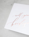 White envelope size card (approx. 3.5" x 5"), reads, "Will you be my bridesmaid?" in rose gold text zoomed in.