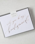 White envelope size card (approx. 3.5" x 5"), reads, "Will you be my bridesmaid?" in rose gold text in a clear plastic bag.