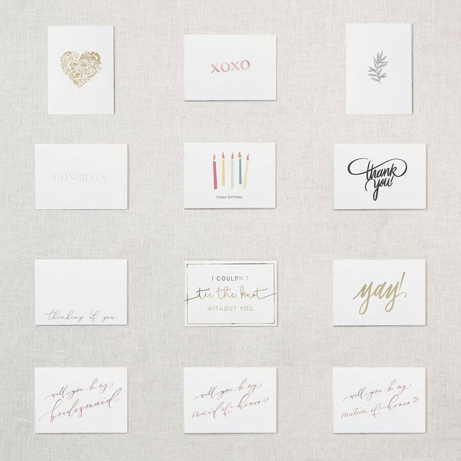 Display of twelve different white cards. Gold foil hear comprised of flowers, Xoxo in rose gold foil, greyscale leafy plant, congrats embossed in white, happy birthday printed underneath five candles of varying colors, Thank You in grey script writing, Thinking of you, I couldn&#39;t tie the know without you in gold, Yay! in gold foil, will you be my bridesmaid in rose gold, will you be my maid of honor in rose gold foil, will you be my matron of honor in rose gold foil.