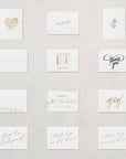 Display of twelve different white cards. Gold foil hear comprised of flowers, Xoxo in rose gold foil, greyscale leafy plant, congrats embossed in white, happy birthday printed underneath five candles of varying colors, Thank You in grey script writing, Thinking of you, I couldn't tie the know without you in gold, Yay! in gold foil, will you be my bridesmaid in rose gold, will you be my maid of honor in rose gold foil, will you be my matron of honor in rose gold foil.