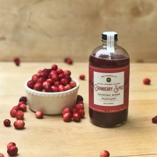 Deep red liquid in a clear glass bottle with a black cap, white seal sticker and red label next to a small bowl of cranberries.