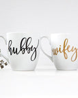 White ceramic mugs, two 15 oz mugs, "hubby" in black script font, "wifey" in gold script font