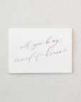 White envelope size card (approx. 3.5" x 5"), reads, "Will you be my maid of honor?" in rose gold text. 