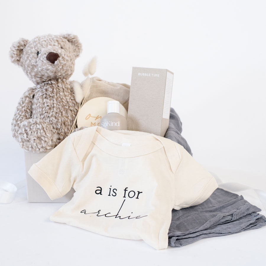 A light grey gift box with SoKind Bubble Time baby wash, a personalized cream onesie with &quot;a is for Archie&quot; printed on it, a charcoal muslin swaddle, wooden monthly milestone baby discs, and Manhattan Toy Co brown Roman bear.