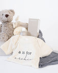 A light grey gift box with SoKind Bubble Time baby wash, a personalized cream onesie with "a is for Archie" printed on it, a charcoal muslin swaddle, wooden monthly milestone baby discs, and Manhattan Toy Co brown Roman bear.