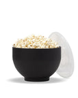 Black silicone popper expanded into bowl holding popcorn with clear plastic lid propped against side. 