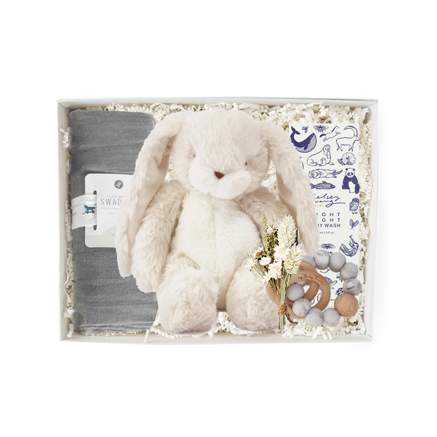 A gray swaddle blanket, a white stuffed bunny, floral bundle, wooden baby rattle, and animal graphic printed box of &#39;night night body wash&#39; in a gray &#39;foxblossom&#39; box with ivory paper shread.